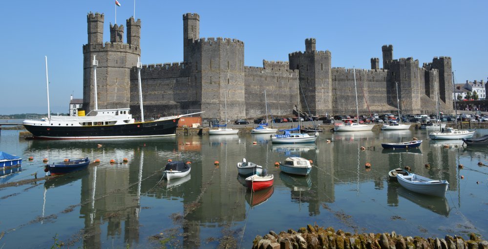 View of the Castle from across the harbour at Caernarfon, with the 3-masted ship BRITAIN berthed on the quayside (2023). She was built in 1910 in Dublin dockyards and was once a lightship, a rig Schooner with motor propulsion (diesel engine) too.  Caernarfon, North Wales